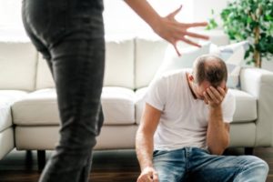Legs of person yelling at man in domestic violence dispute while man sits on floor crying.