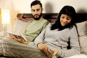 A couple reading mobile phone and tablet in bed and man is concerned about cyberspying and divorce.