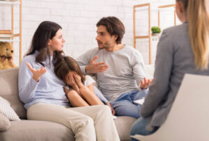 Parents arguing during a parenting coordinator session, with a child visibly distressed between them. A professional mediator is present, guiding the conversation.